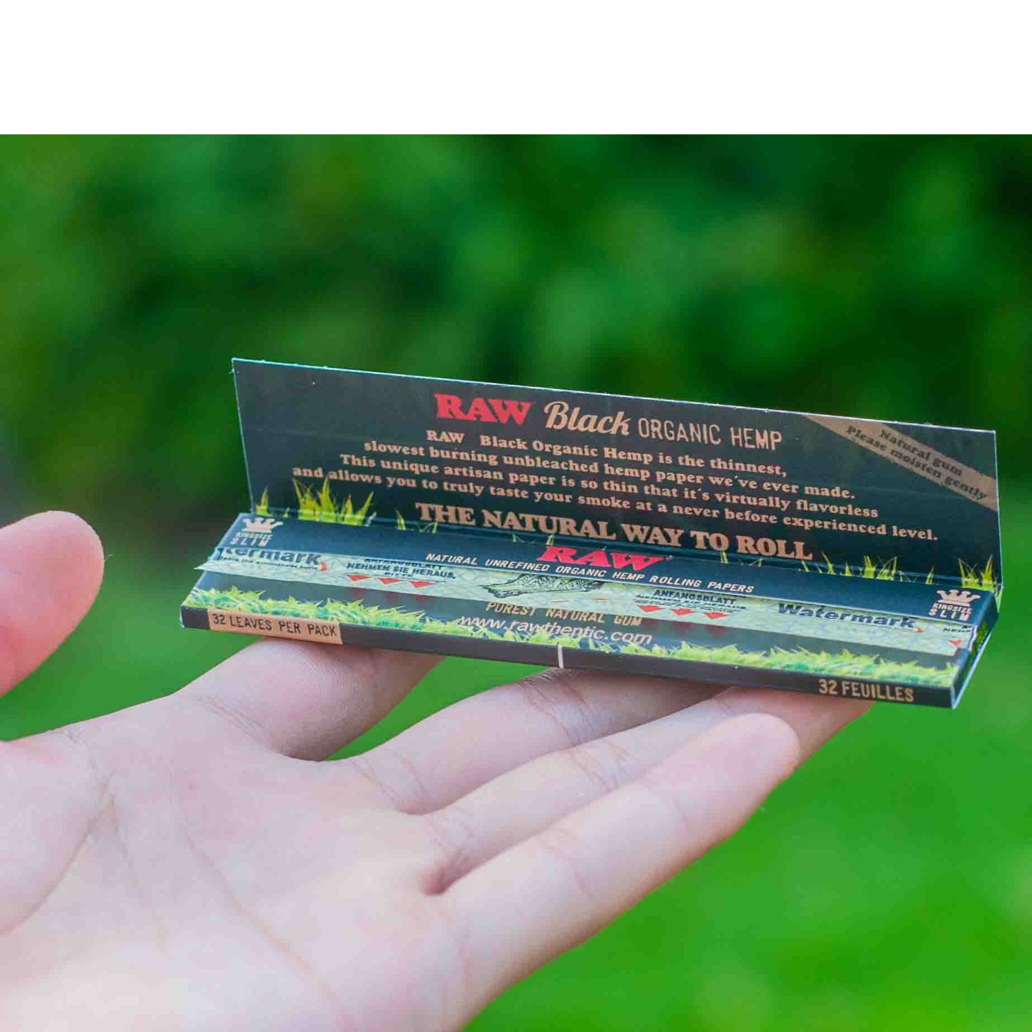 Hand holding a pack of RAW Black Organic Hemp rolling papers against a green background